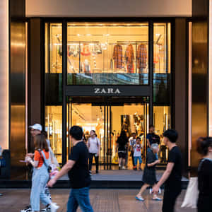 人們經過一間有玻璃窗和溫暖室內照明的Zara服裝店。 — Zara 壁紙