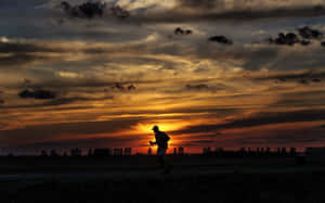 夕陽下田野上跑者的剪影，伴隨著戲劇性的橙色雲彩。 — 日落 壁紙