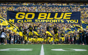足球隊在球場上慶祝，球迷揮舞旗幟並舉著「Go Blue」橫幅。 — 密西根大學橄欖球 壁紙