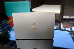 戶外玻璃桌上的銀色MacBook，背景有庭院家具。 — 筆電 壁紙
