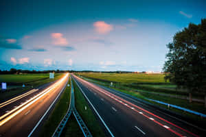 黃昏時分的高速公路，帶有流線型汽車燈光和兩旁的綠色田野。 — 藍天 壁紙