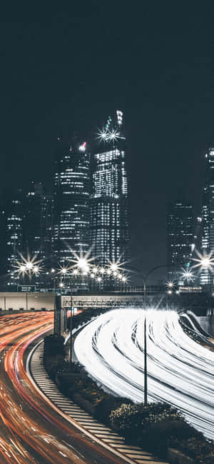 夜間城市景觀，在發光的摩天大樓下彎曲的高速公路上的汽車燈光條紋。 — iPhone 17e 壁紙