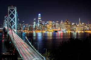 夜間橋上燈光軌跡橫跨水面，背景是發光的城市天際線。 — 舊金山 壁紙