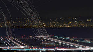 夜間機場跑道，帶有飛機燈光軌跡和遠方城市光芒。 — 伸展台 壁紙
