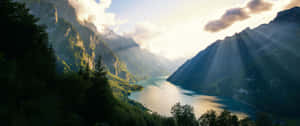 陽光穿過雲層，照在一個被綠色山峰和樹木環繞的山間湖泊上。 — 湖泊 壁紙