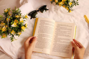 雙手正在閱讀一本書，附近有黃色花朵和太陽眼鏡。 — 花朵 壁紙
