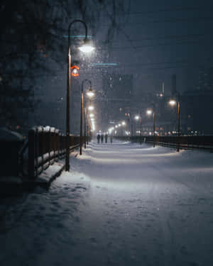 雪落在燈火通明的城市步道上，遠處有人行走。 — 夜晚 壁紙