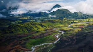 一條蜿蜒的河流流經鬱鬱蔥蔥的綠色和棕色火山山谷，背景是山脈。 — 冰島 壁紙