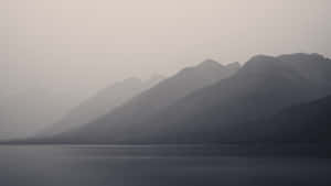 在朦朧的黎明天空下，霧氣籠罩的山峰映襯著寧靜的山間湖泊。 — 山景 壁紙
