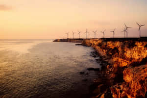 夕陽下懸崖上的風力發電機，將溫暖的光線投射在海洋和岩石上。 — MacBook Air M2 壁紙