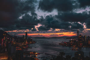 黃昏時分的香港天際線，城市燈光與港灣水面在戲劇性的雲層下。 — 城市 壁紙
