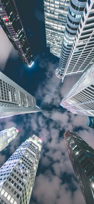 夜間向上看，高聳的照明摩天大樓映襯著多雲的天空。 — 1080x2340 壁紙
