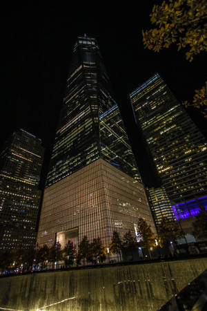 夜晚的城市景觀，有燈火通明的摩天大樓和秋樹，靠近反射的水景。 — 小米平板 9 Pro 壁紙