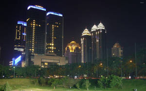 夜晚城市天際線，摩天大樓燈火通明，前景是綠色公園 — 印尼 壁紙