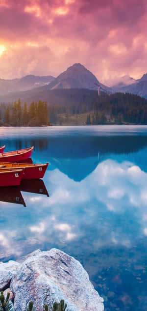 紅色獨木舟漂浮在平靜的湖面上，反射出山脈和粉紅色的日落雲彩。 — Pixel 6 Pro 壁紙