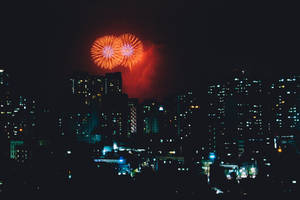 煙火在夜間城市上空綻放，下方是燈火通明的建築物。 — 城市 壁紙