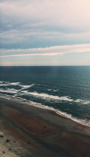 鳥瞰海洋波浪在沙灘上拍打，天空多雲。 — 海灘 iPhone 壁紙