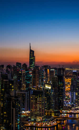 黃昏時分的現代城市天際線，燈火通明的建築物和閃耀的天空倒映在水中。 — Galaxy Tab A 壁紙