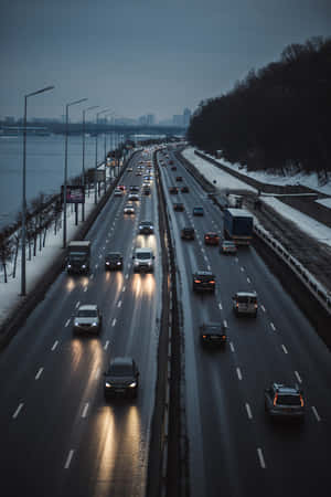 積雪的高速公路，汽車在黃昏時分行駛，靠近河流和城市天際線。 — 2:3 壁紙