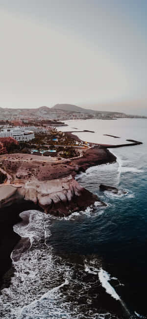 黃昏時分的沿海小鎮，海浪拍打著黑暗的懸崖，建築物沿著海岸排列。 — iPhone 18 壁紙