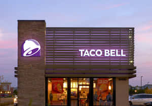 黃昏時分Taco Bell餐廳外觀，帶有發光招牌和溫暖的室內燈光。 — 塔可 壁紙