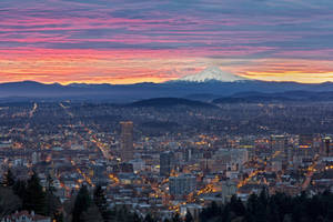 城市燈光在 vibrant 的日落天空下閃爍，遠處有白雪覆蓋的山峰。 — 俄勒岡州 壁紙