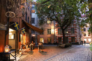黃昏時分的鵝卵石庭院咖啡館，帶有瑞典國旗和溫暖燈光。 — 暮光 壁紙