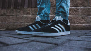 黑色Adidas Gazelle運動鞋帶白色條紋和藍色牛仔褲，在磚砌路面上。 — 愛迪達 GIF 壁紙